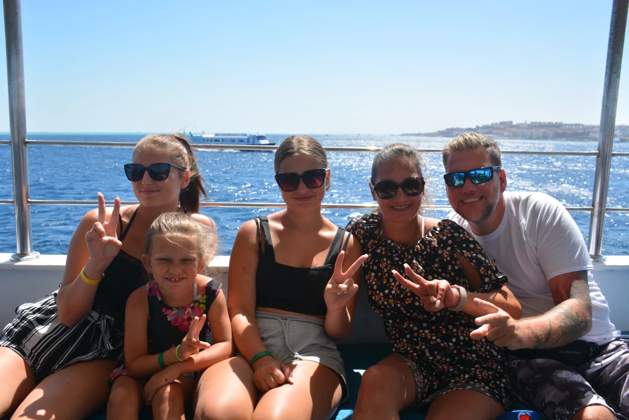Fünfköpfige Familie mit Sonnenbrillen auf dem Deck eines Ausflugsboots, dahinter das Rote Meer, ein weiteres Schiff und die Küste von Hurghada